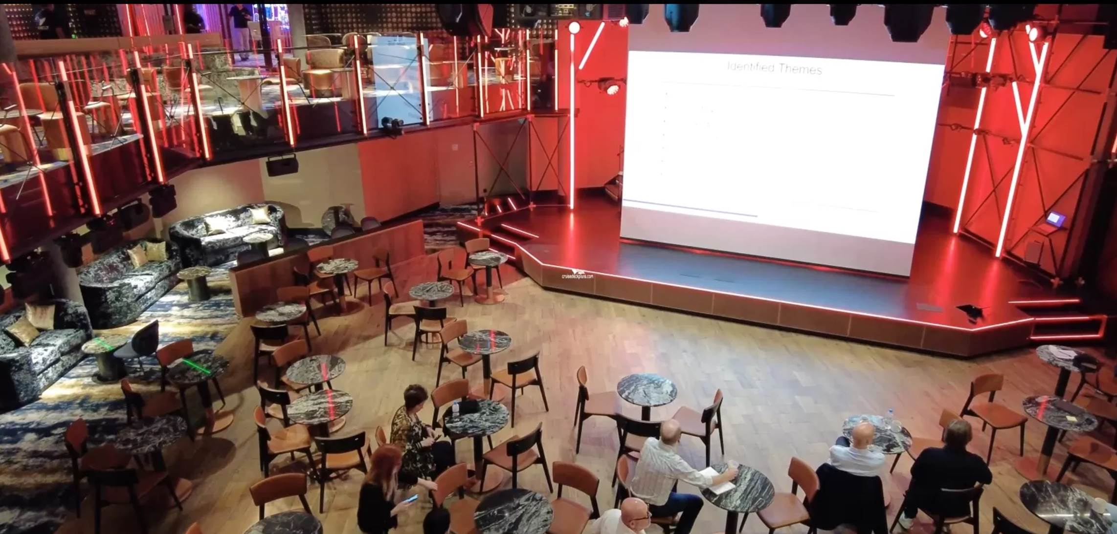 A group of people sitting at tables and chairs in a room with a projector screen

Description automatically generated